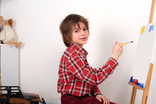 der Junge im roten Hemd in einem Ausschnitt zeichnet Farben auf eine Staffelei - Foto, Bild