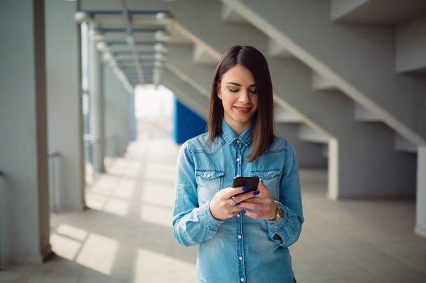 Приваблива молода дівчина в сорочці надсилає повідомлення через свій смартфон, стоячи в коридорі
 - Фото, зображення