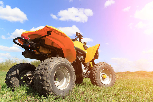 A four-wheeled yellow ATV quad-bike standing idle on the green grass, with trees and a blue sky with clouds on the background - 写真・画像