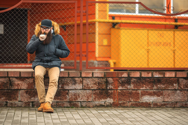 Vape bearded man in real life. Portrait of young guy with large beard in glasses and a cap vaping an electronic cigarette opposite the fence in the - Valokuva, kuva
