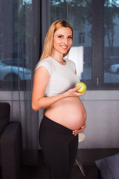 Belleza saludable. Hermosa joven embarazada sosteniendo manzana verde y mirando a la cámara con sonrisa mientras está de pie en el interior del hogar
 - Foto, Imagen