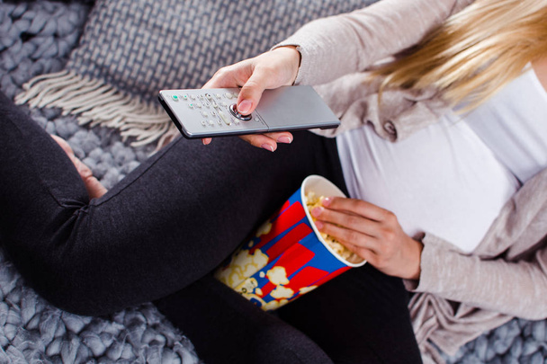 Время смотреть телевизор! Крупным планом молодая беременная женщина смотрит телевизор и ест попкорн, сидя на диване в интерьере дома
 - Фото, изображение