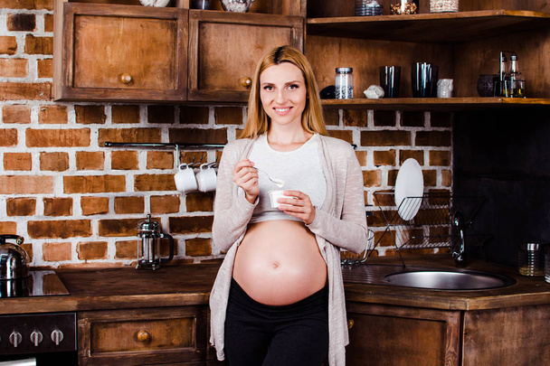 Joghurt zum Mittagessen? schöne junge schwangere Frau öffnet Kühlschrank und blickt lächelnd in die Kamera, während sie im Kücheninneren steht - Foto, Bild