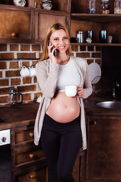 Увидимся завтра? Красивая молодая беременная женщина разговаривает по телефону и смотрит в камеру с улыбкой, стоя на кухне интерьера
 - Фото, изображение