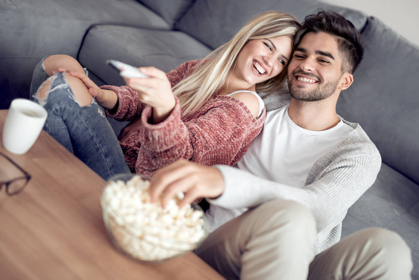 Νεαρό ζευγάρι χαλαρώνοντας στο διαμέρισμά τους, κάθεται στον καναπέ, βλέποντας μια ταινία και τρώγοντας ποπ κορν. - Φωτογραφία, εικόνα