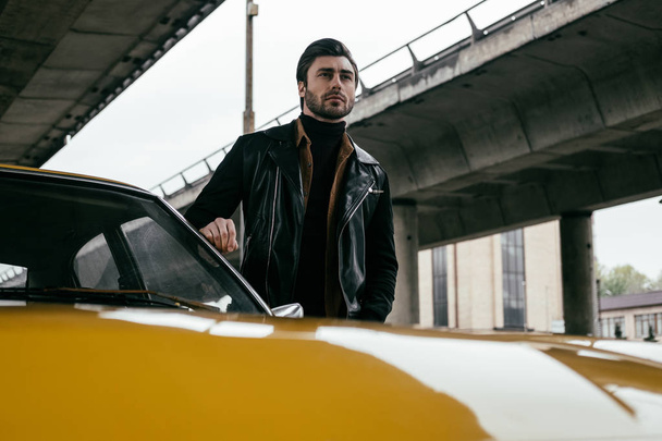 красивий стильний молодий чоловік в шкіряній куртці стоїть біля жовтого класичного автомобіля і дивиться в сторону
 - Фото, зображення