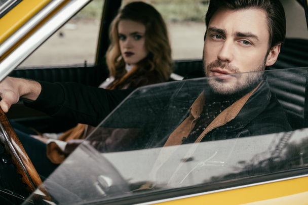 pohledný elegantní mladý muž, zatímco sedí v autě s krásnou přítelkyní při pohledu na fotoaparát - Fotografie, Obrázek