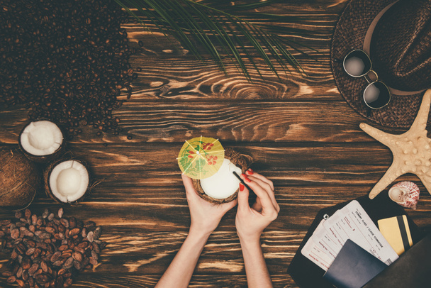 tiro recortado de mulher com coquetel de coco cercado com vários atributos de viagem tropical na superfície de madeira
 - Foto, Imagem