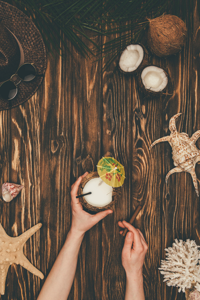 cropped shot of woman with coconut cocktail and cigar on wooden surface surrounded with various tropical travel attributes - Photo, Image