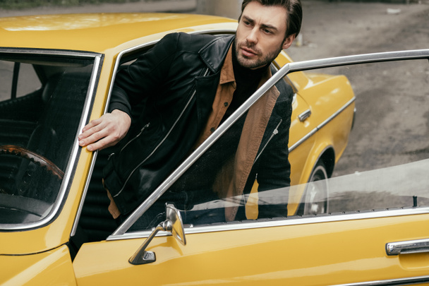 handsome stylish young man in leather jacket opening door of yellow classic car and looking away - 写真・画像