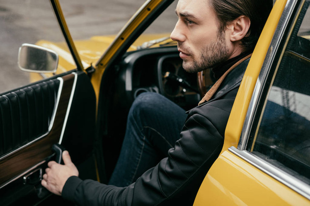 side view of handsome stylish man opening door and looking away while sitting in retro car - 写真・画像