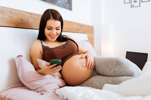 Pregnant woman using phone while lying on bed and resting - Foto, Imagem