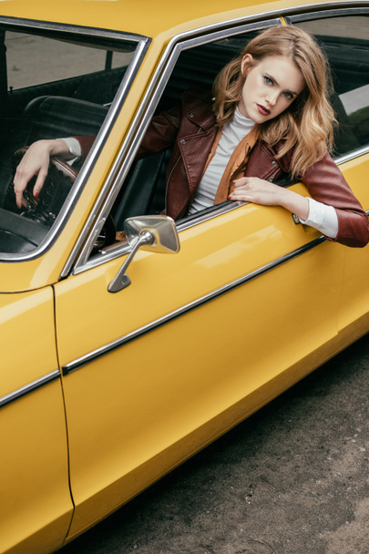 beautiful stylish girl looking at camera through window of yellow retro car - Foto, afbeelding