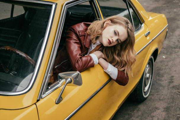 high angle view of beautiful stylish girl looking at camera through window of yellow classic car  - Foto, imagen
