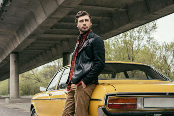 стомлений стильний чоловік у шкіряній куртці, що стоїть біля старовинного автомобіля і дивиться геть
 - Фото, зображення