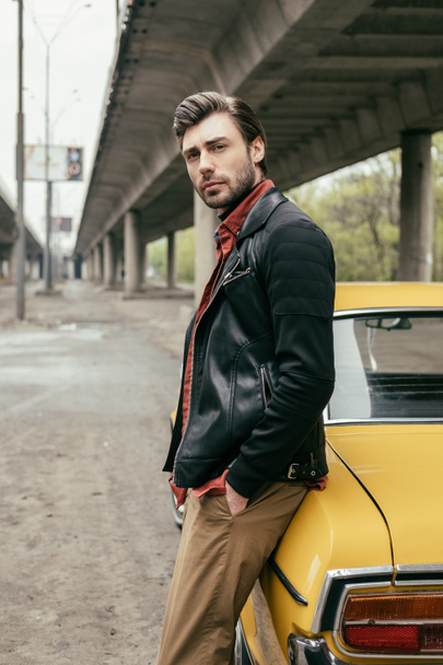 handsome stylish man in leather jacket leaning at yellow car and looking at camera - Photo, Image