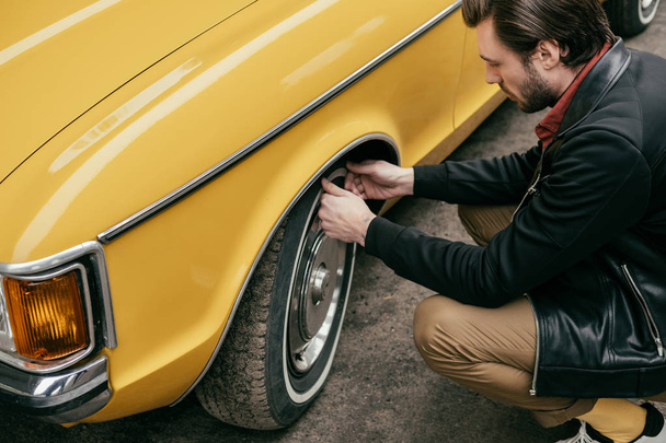 cropped shot of handsome man in leather jacket fixing wheel of yellow retro car  - Photo, Image