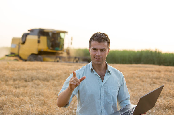 Ingeniero joven agricultor feliz con portátil de pie en el campo de trigo y apuntando con el dedo a la cámara mientras combina la cosechadora trabajando en segundo plano
 - Foto, imagen