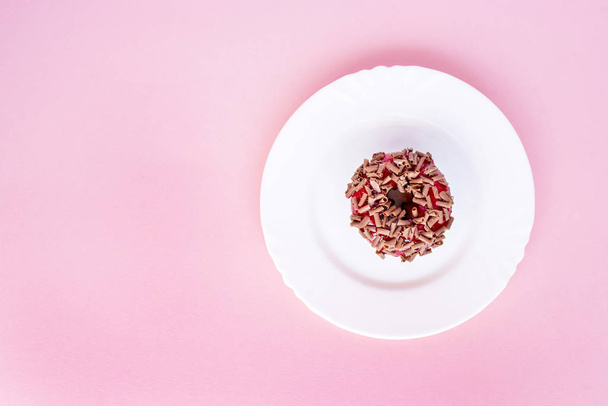 Пончик покрыт красной глазурью и посыпан тертым шоколадом на белой тарелке на розовом фоне
 - Фото, изображение