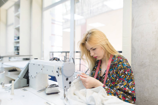 Una costurera linda trabaja en el lugar de trabajo cerca de una máquina de coser. Lugar de trabajo para costureras. La chica está cosiendo su ropa en casa
 - Foto, Imagen