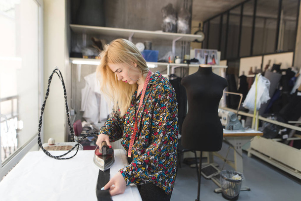 A seamstress girl is standing in a studio near an ironing board and ironing clothes. Workplace. Creation of fashion clothes in the studio - Photo, Image