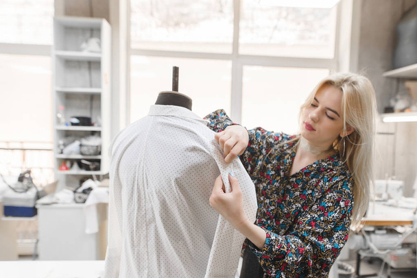 Young female dressmaker works inserts needles into the clothes that are on the mannequin. Fashion designer creates stylish clothes in the studio. Fashion designer concept. Focus on clothes - Foto, Imagem
