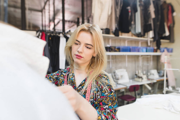 Portret van een jonge mode-ontwerper op het werk. Jonge naaister vrouw staat in een studio in de buurt van een manicure met kleding en werken. Stijlvolle kleding maken - Foto, afbeelding