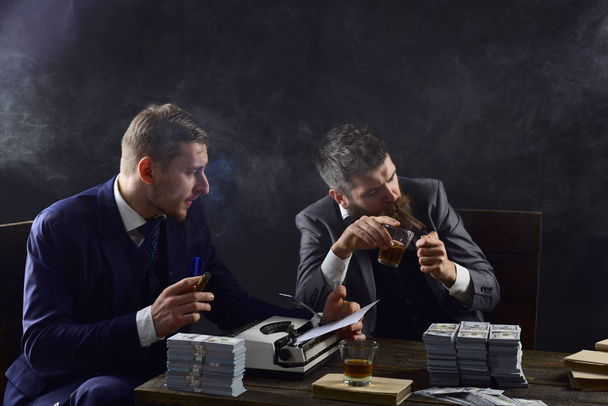 Men sitting at table with piles of money and typewriter. Illegal business concept. Businessmen discussing illegal deal while drinking and smoking, dark background. Company engaged in illegal business. - Photo, image
