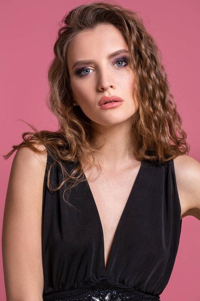 Beautiful fashionable girl with long curly hair in a black dress in the studio on a pink background. - Fotoğraf, Görsel
