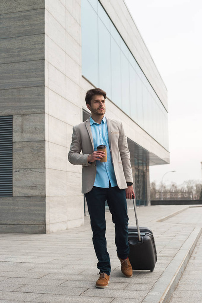 Тільки вперед! Фотографія молодого красивого підприємця, який витягує маленьку багажну валізу з будівлі аеропорту і п'є каву після прибуття
. - Фото, зображення