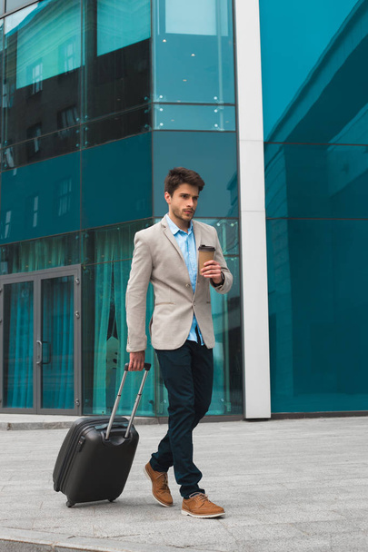 Success is often achieved! Full length vertical shot of young handsome male business traveler pulling baggage on city street, while drinking coffee. - Photo, Image