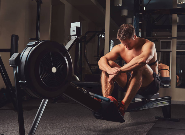 Atleta musculoso sin camisa descansando después de un duro entrenamiento mientras se sienta en el remo en el gimnasio
. - Foto, imagen