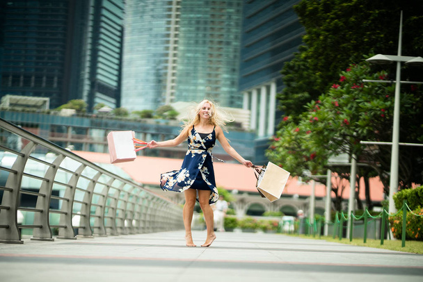 Η χαρά του ψώνια... Ενθουσιασμένος όμορφη γυναίκα που φοράει περιστασιακή μπλε φόρεμα με λουλούδια, τσάντες για ψώνια στους δρόμους με ουρανοξύστες  - Φωτογραφία, εικόνα