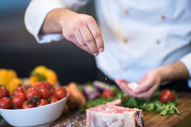 Master Chef hands putting salt on juicy slice of raw steak with vegetables around on a wooden table - Fotó, kép