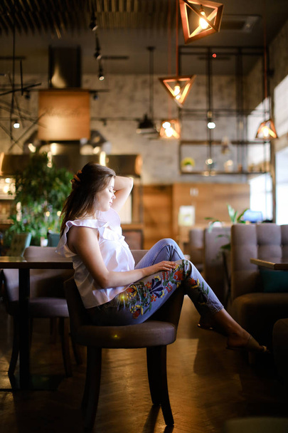 Vrouwelijke foto-model zit op café met voeten op achterzijde stoel, dragen van etnische jeans. - Foto, afbeelding