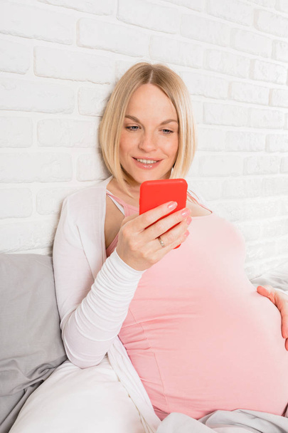 Happy smiling pregnant woman lies in bed and looks at mobile phone - 写真・画像
