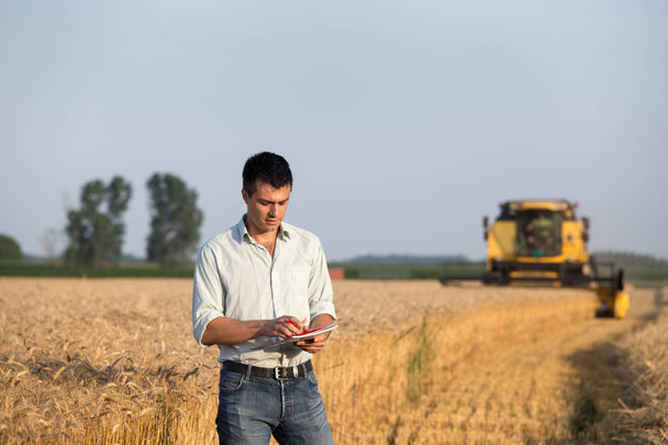 Young farmer engineer with notebook standing on wheat field while combine harvester working in background - Photo, Image