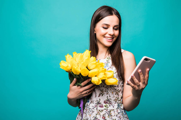 Portrait of a happy young girl in dress use mobile phone while holding big bouquet of yellow tulips isolated over color background - Photo, image