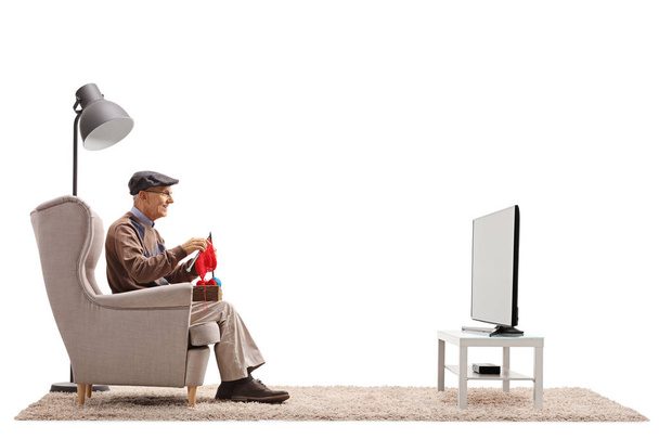 Elderly man seated in an armchair knitting and watching television isolated on white background - Фото, изображение