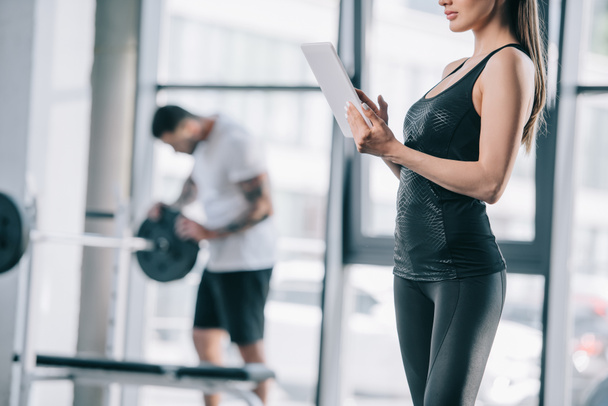 körülvágott kép női személyi edző, digitális tabletta és sportos férfi súlyzó-on tornaterem - Fotó, kép