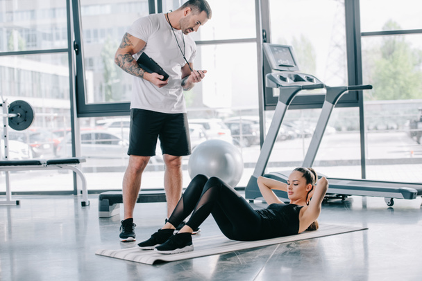 male personal trainer with timer and athletic woman doing abs on fitness mat at gym - Foto, Imagem