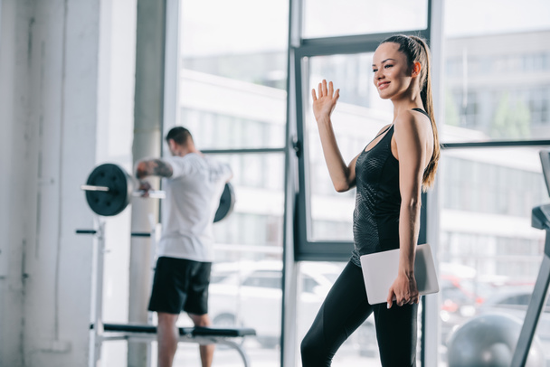 női személyi edző, digitális tabletta integetett kézzel és sportos férfi súlyzó mögé edzőteremben gyakorlása - Fotó, kép