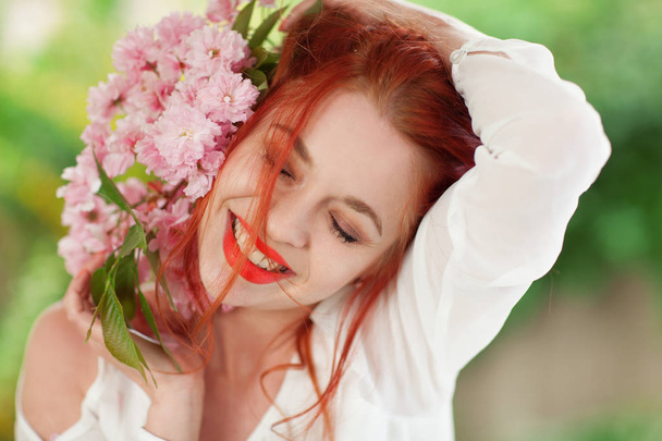 Beautiful young woman with red hair having fun standing in the garden with cherry blossom branch and perfect make up - Photo, image