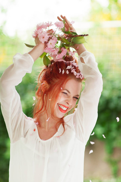 Beautiful young woman with red hair having fun standing in the garden with cherry blossom branch and perfect make up - Фото, зображення
