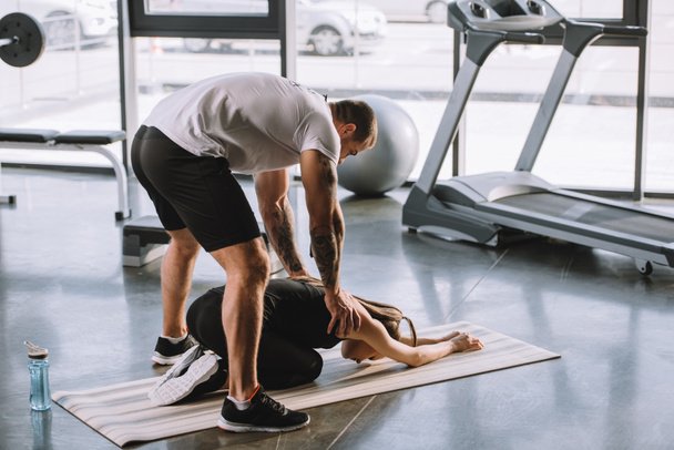male personal trainer helping young athletic woman to stretching on fitness mat at gym - Photo, image