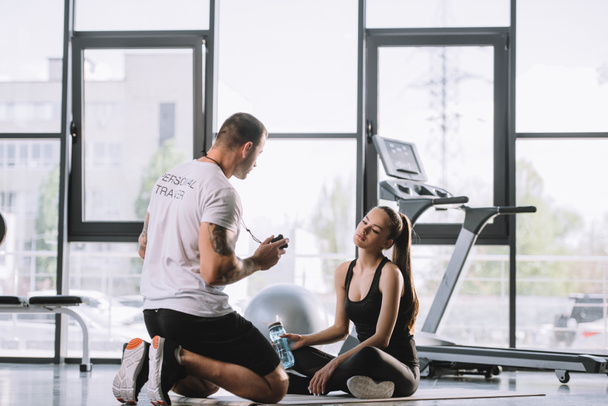 male personal trainer showing timer to young athletic woman sitting on fitness mat with bottle of water at gym - Fotoğraf, Görsel