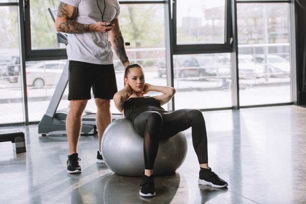 mannelijke persoonlijke trainer met timer en jonge sportieve vrouw doet abs op fitness bal op sportschool - Foto, afbeelding