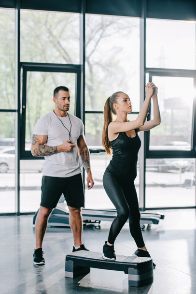 junge Sportlerin macht Step-Aerobic und männlicher Personal Trainer mit Zeitschaltuhr in der Hand im Fitnessstudio - Foto, Bild