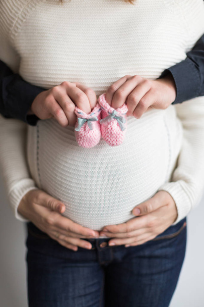 baby fashion, love, pregnancy concept. there is close up of female and male arms, young mother is holding her hands on her belly and her husband is holding a couple of cute pink baby bootees - Photo, Image