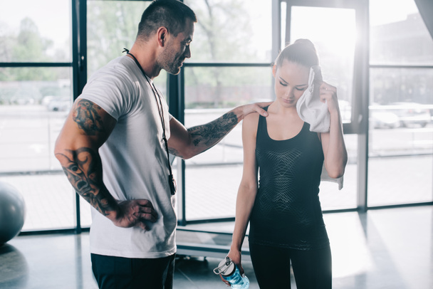 male personal trainer cheering up young sportswoman with bottle of water wiping face with towel at gym - Foto, Imagem